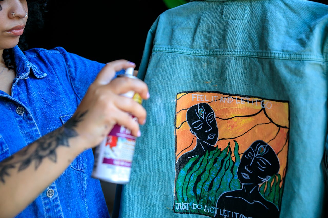 gallery-6 Woman artist spray painting a denim jacket with unique artwork design.