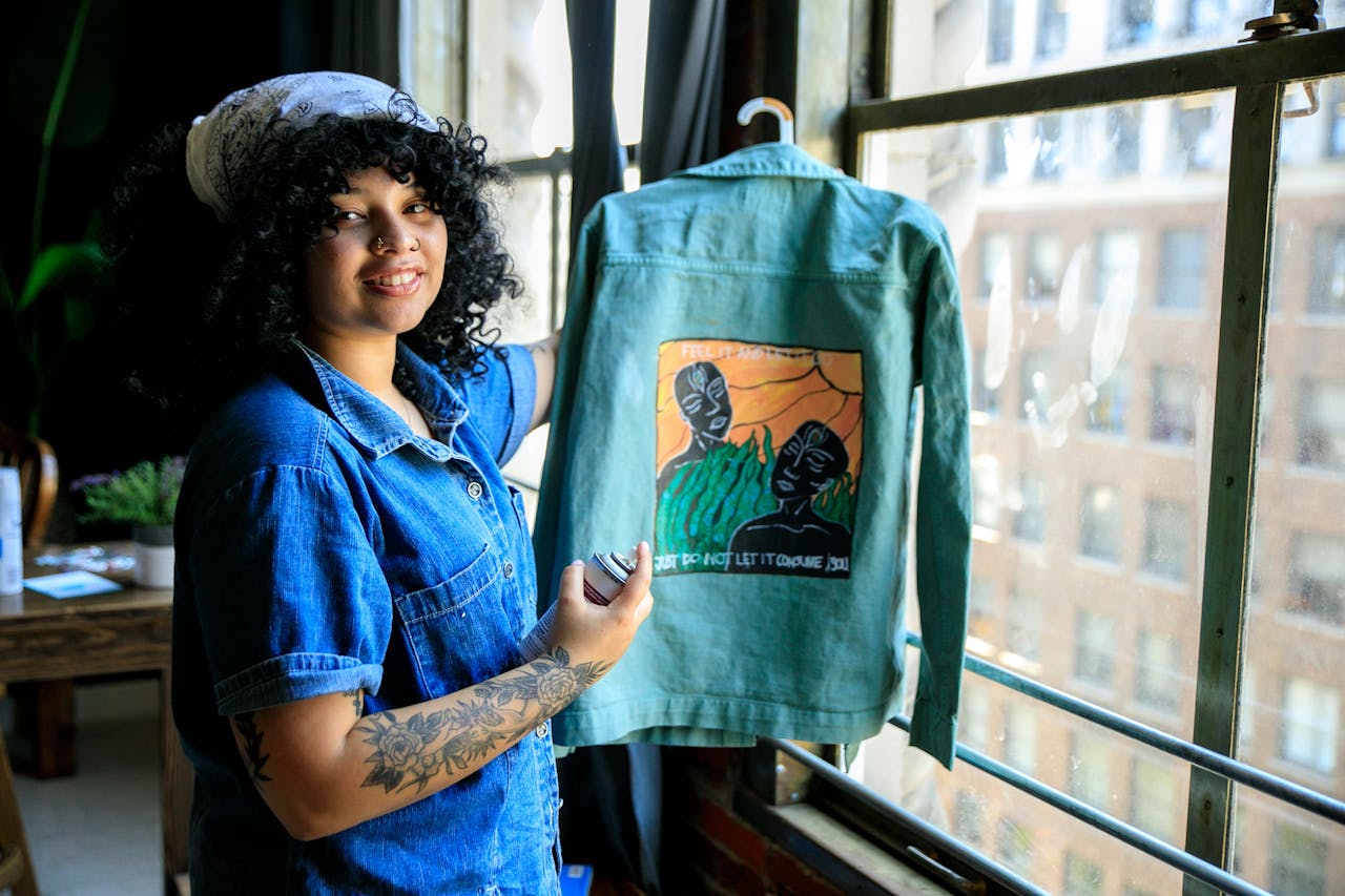 hero-img-01 Smiling woman with tattoos displays a custom painted jacket by a window indoors.