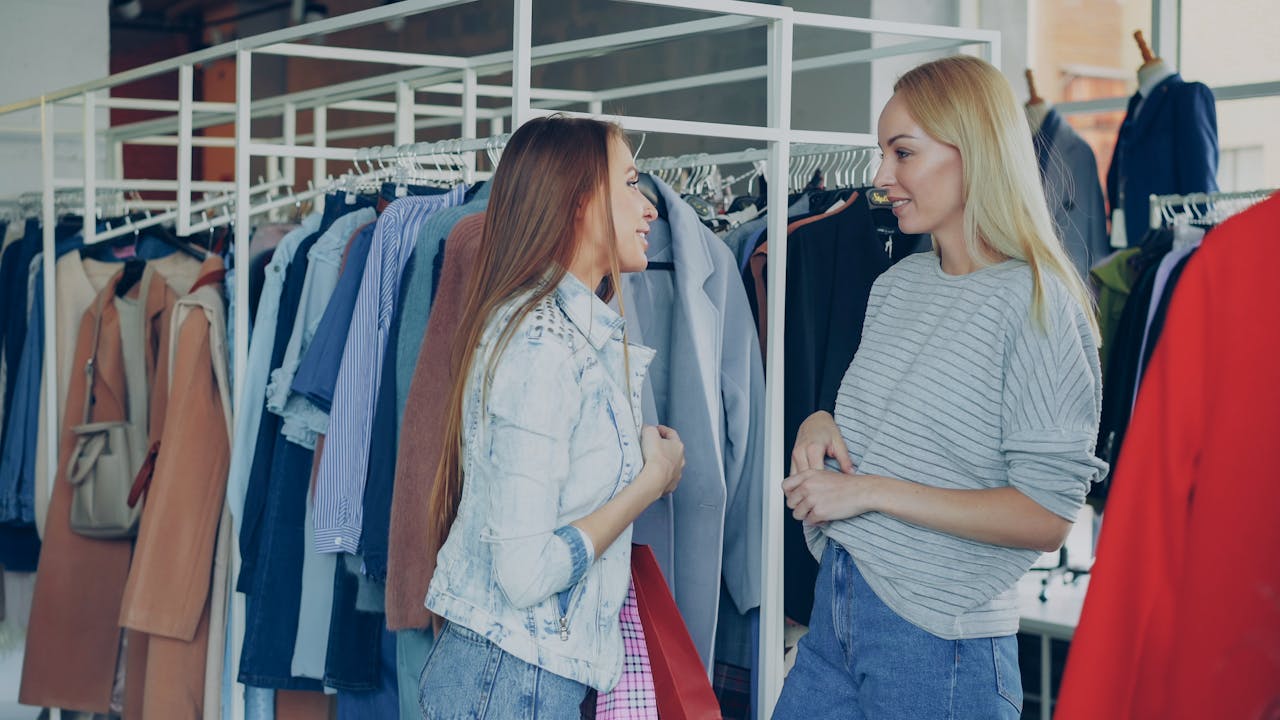 hero-img-02 Two women chatting among clothing racks in a stylish fashion store, sharing shopping advice.